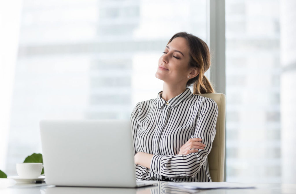 A woman sitting in an office chair relaxing with her eyes closed, taking a break from working on her computer to avoid digital eye strain.