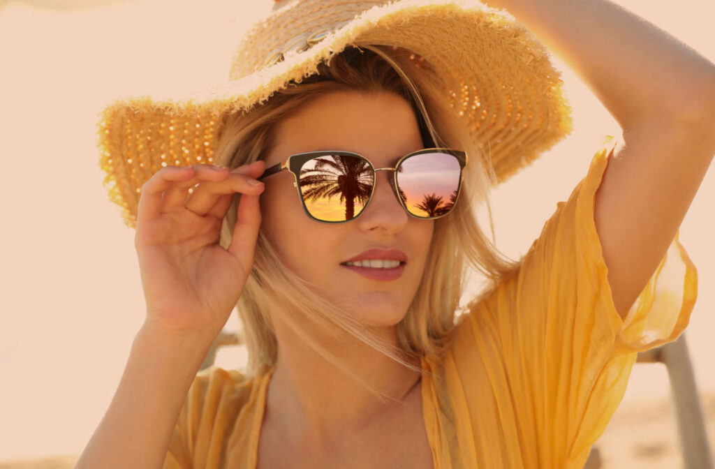 A beautiful young woman wearing polarized sunglasses and a straw hat at a beach in a sun-glazed yellow background.