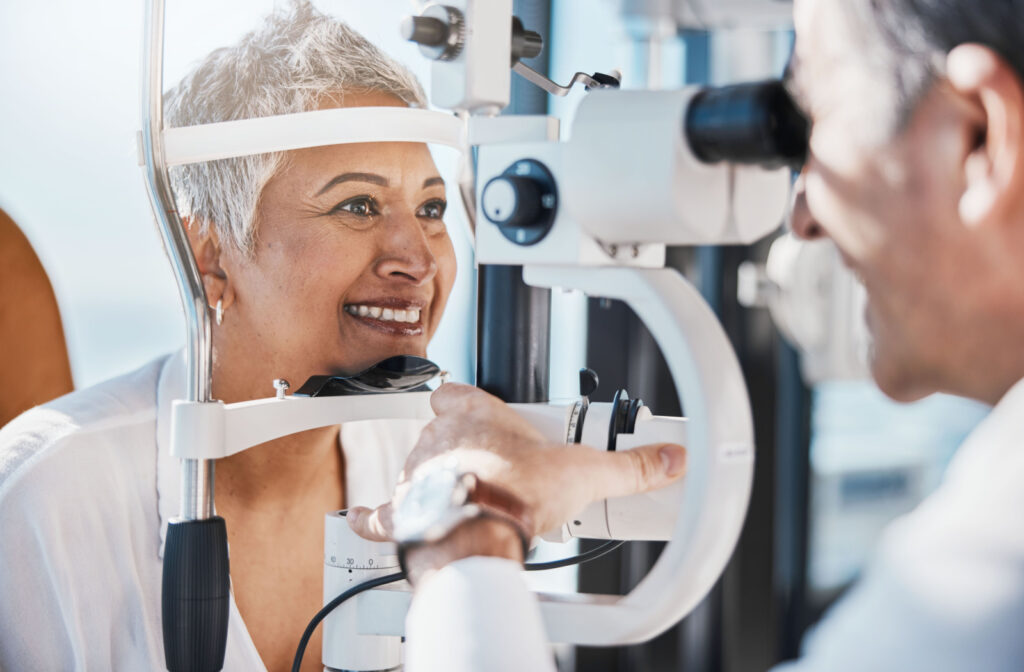 A woman smiles while she rests her chin and forehead on a machine and a male eye doctor performs an exam.