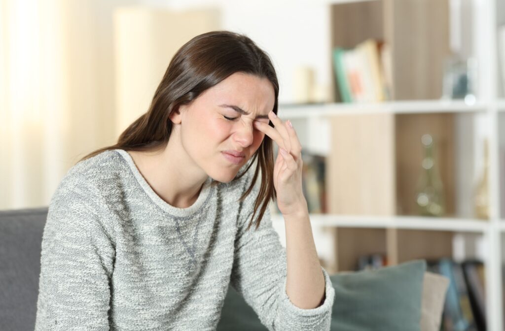 A young adult sitting at home, rubbing their eyes and squinting in frustration due to burning feelings from their contact lenses.