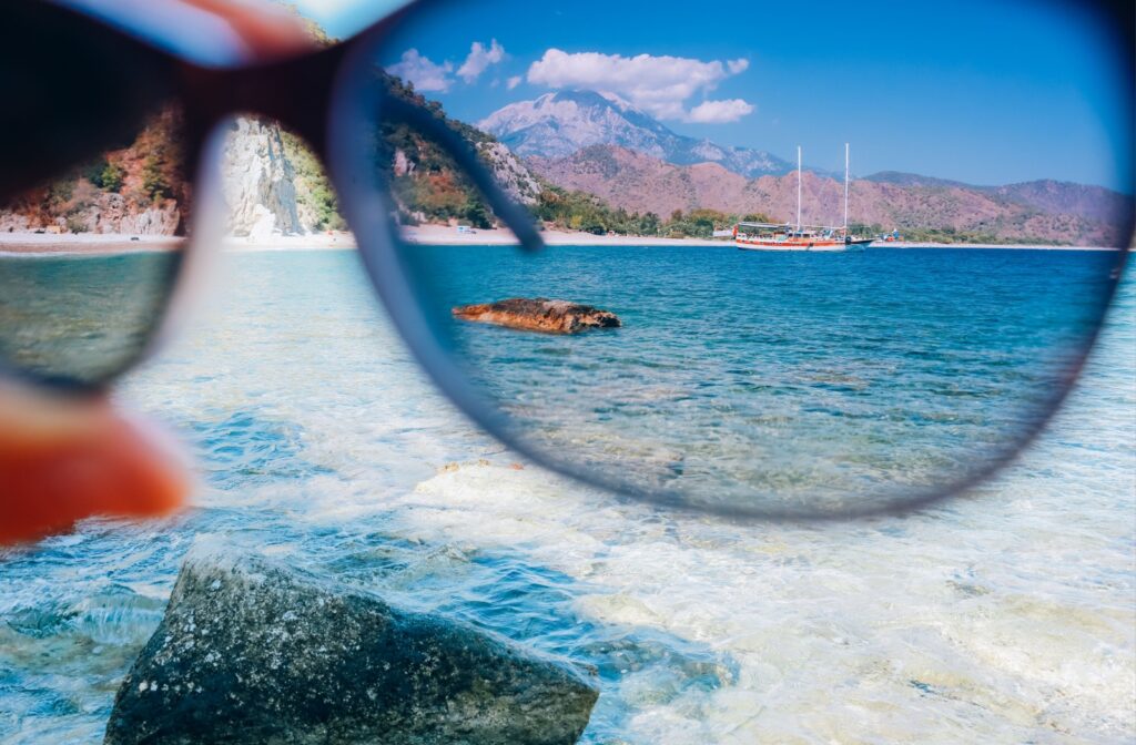 A hand holds polarized sunglasses against a body of water, showing how vibrant and sharp vision becomes.